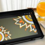 A tablet displaying mandala artwork beside a glass of orange juice on a table.